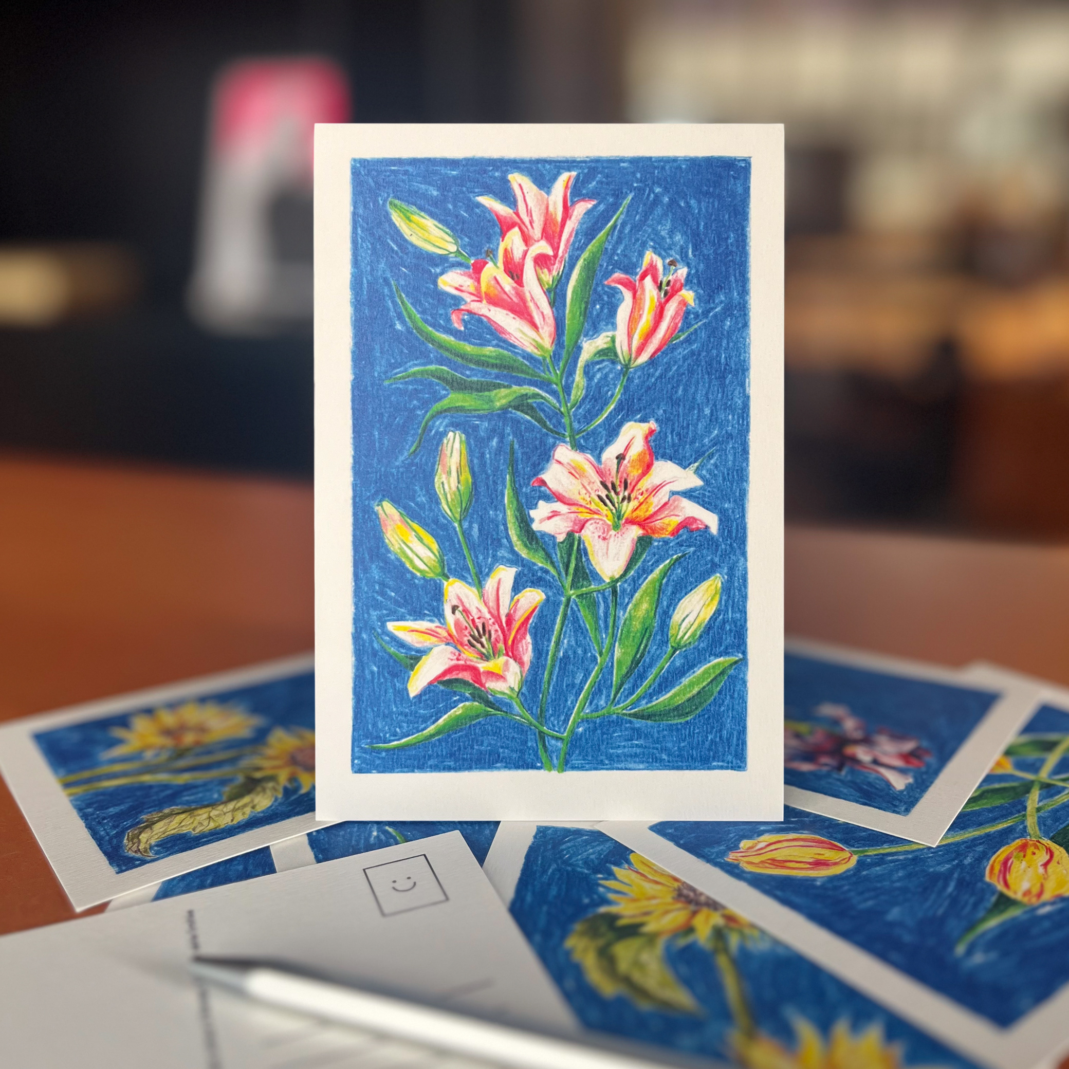 Carte postale illustrée de lys au crayon de couleur par Yollema, posée sur un bureau avec d'autres cartes postales