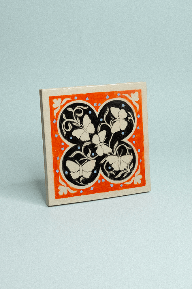 Carreau de grès décoratif, peint à la main, avec cinq papillons beiges sur fond noir et orange avec un motif floral symétrique.