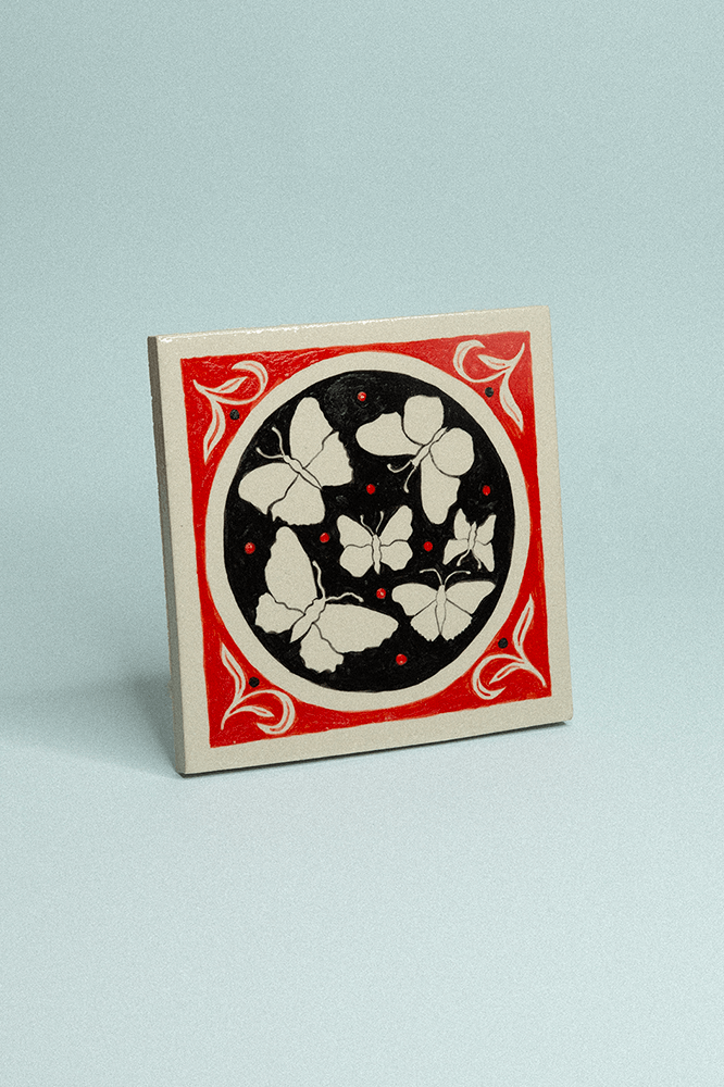 Carreau de grès décoratif, peint à la main, avec six papillons beiges sur fond noir et rouge.